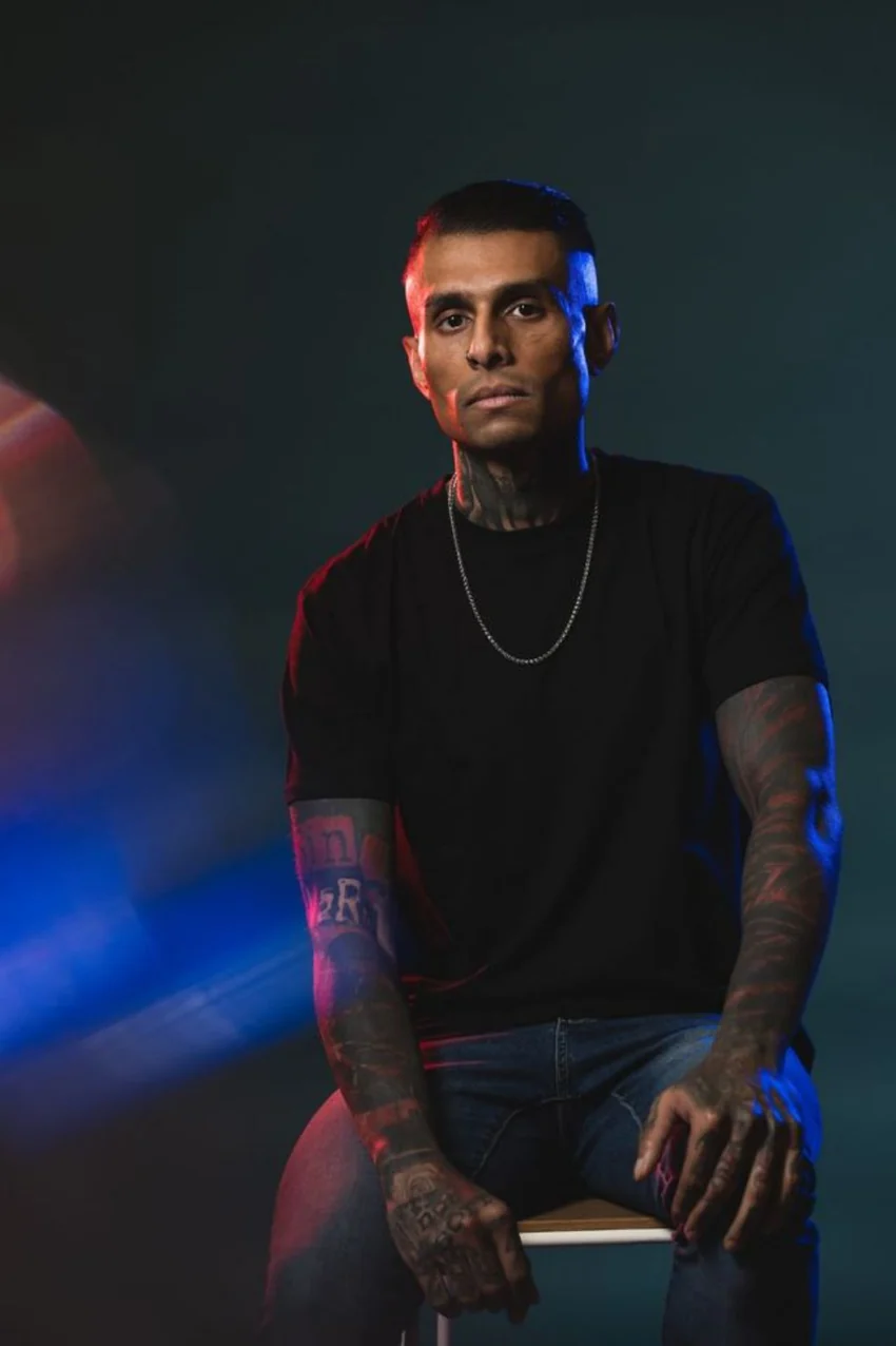 Shefan, the creator of Raw By Design, seated in a black T-shirt under red and blue editorial lighting